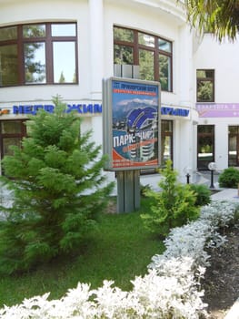 advertising board on a green flowerbed with trees, flowers and lanterns in front of a modern white building