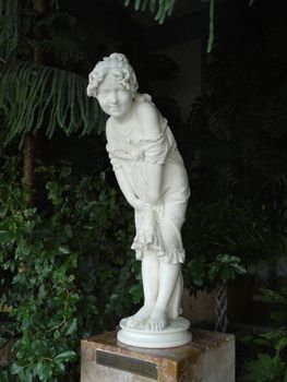 The sculpture of a woman is bashfully covered with clothes made of gypsum. Standing on a low stand next to the green shoots of plants.