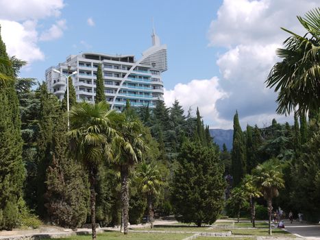 A gray skyscraper in an oasis of palm trees and other trees against a blue sky with celestial clouds