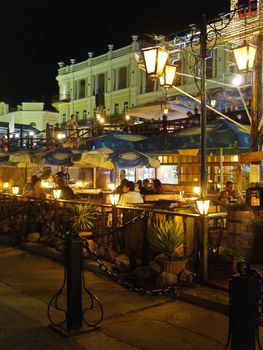 A city evening with people having a rest at tables of a cafe in the open air with a lantern near the table creating a romantic atmosphere and live music.