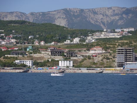 Boat sailing in the sea near the shore with tourists on holiday with the houses of the spa town seen on the coast against the backdrop of high mountains on the horizon.