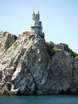 A low house with a tower located at the very edge of the cliff and tourists looking at the beauty of the sea from the balcony