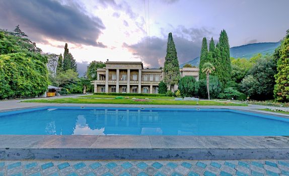 chic light two-storey villa with a lawn, a large blue pool surrounded by tall green trees against the backdrop of the mountains and the gloomy sky. place of rest, travel, tourism
