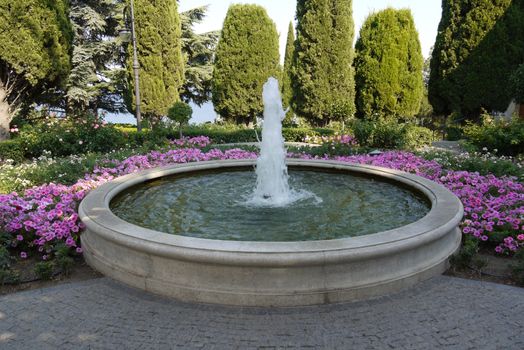 A beautiful little fountain in a huge bowl of water with flowers growing around it against the background of trees in the distance.