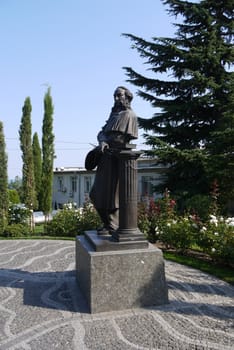 monument to a man of the 18-19th centuries on the square in front of the building, surrounded by flowering flower beds and tall green trees under a blue cloudless sky