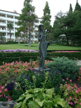 a statue of a beautiful girl in the middle of a flower bed amid tall green trees and a white hotel building, a recreation center, a sanatorium with large balconies. place of rest, travel, tourism