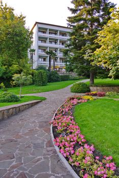 A stone path with planted flowers, tall green trees and a lawn leads to a white five-story building with balconies. place of rest, hotel, hotel, sanatorium