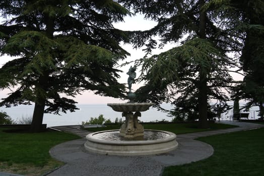 decorative fountain with statue of a girl on the lawn surrounded by fir trees against the background of the sea and sky. place of rest and travel