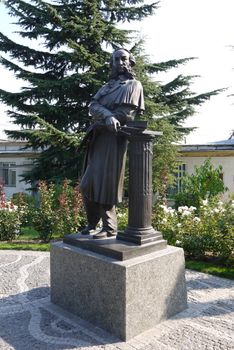 A monument with a standing man leaning on a column with large sideburns and drawing brushes. Located next to the flowerbed with flowers and a high lush spruce.