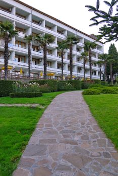 stone path through a green lawn to a beautiful hotel with a cafe on the terrace under umbrellas