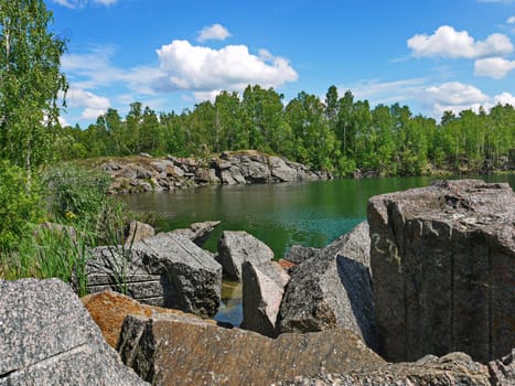 Deep green lake with large stone boulders and trees on the sides against a blue sky with white clouds