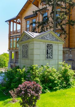 a small decorative building of white stone in front of a huge wooden mansion on a background of bright juicy greens and a blue sky