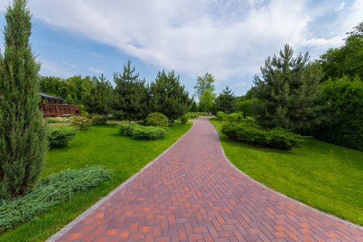 The path in the park is laid out with red tiles between green grasses and ornamental bushes. Mezhygirya Ukraine