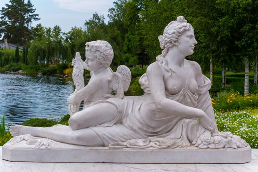 Beautiful sculpture of a half-naked woman with a little boy with wings behind her back against the backdrop of a pond in a park of flowers and green nature