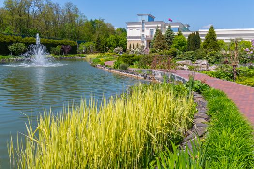 The scenery in the park is a bright sunny day with the magnificence of green plantings of flowers by a pond with a fountain of tall beautiful trees and a light building with a flying flag on the roof.