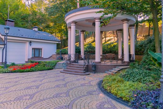 Spacious gazebo with marble floor and steps against the background of green flower beds