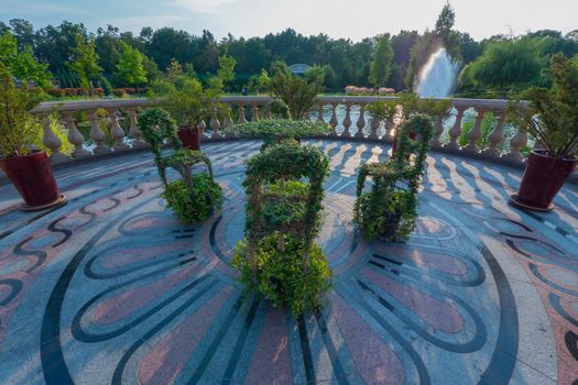 A chic composition of chairs and a table panned by green plants standing on a round platform paved with patterned tiles with a beautiful view of the pond with a fountain.