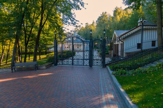 Entrance to the territory closed with an iron gate with a door made of their figured lattices with lanterns on top with a bench beside him in the shade of trees.