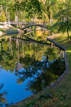A small bridge across a pond with a quiet smooth surface in which, as if in a mirror, there are trees and a piece of blue sky standing on the shore.