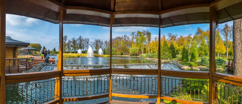 Luxurious view from the gazebo to relax on a pond with fountains and resting people nursing small ducklings.