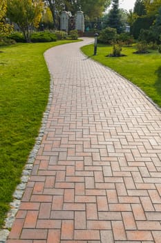 A paved path between green lawns with bushes and trees going to the gate with a lattice fence standing away.