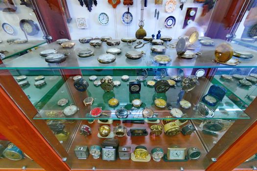 Antique glass showcase with a lot of different clock movements, as well as a hanging clock in the background