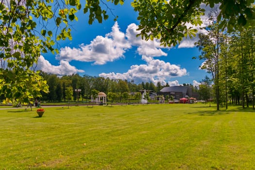 A bright green lawn in the park on a sunny clear day with a covered pavilion in the distance, against a bright blue sky with white clouds.