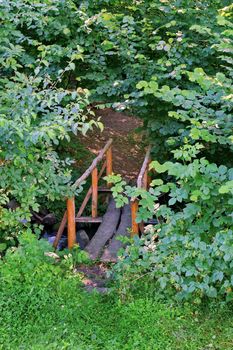 A wooden bridge made by hand lying over a shallow stream with stones running in the forest among the trees.
