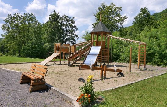 Children's playground covered with sand with a slide for a swing with a wooden deck standing bench for rest and a growing bush color next to it.