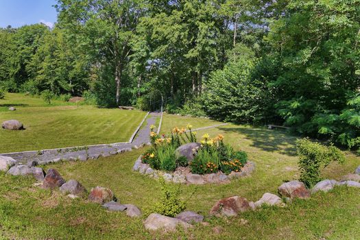 A small bench standing near thick bushes on a green lawn with a beautiful flower bed with flowers surrounded by stones next to it.