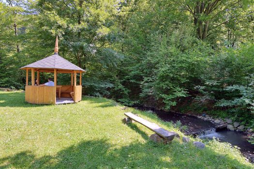 gazebo and bench for relaxation on a green carpet of grass on the bank of a forest river
