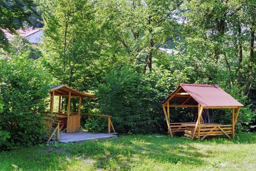 A shelter for rest with benches and a table standing in the shade of bushes and trees on the green grass. A good place to relax in peace and solitude.