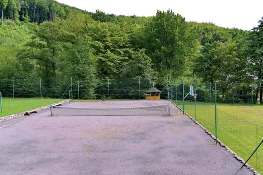 A tennis court with a stretched mesh in the middle and a number of sports grounds for playing basketball and football on the background of green trees behind the fence net.