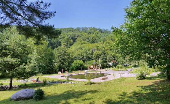 Clean pool with wooden benches, sun loungers and a playground in the background of the park zone