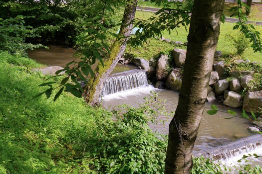 small waterfalls of running water through logs in a shallow river with a bunch of stones on the shore