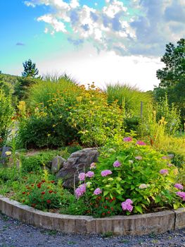 A colorful flowerbed with bright flowers and green grass with large stones in it surrounded by a curb. Located against the background of this sky with clouds.