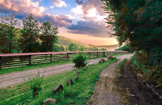 The dusty road is enclosed by a small fence against the background of a beautiful sky with divorces. The clouds play in all shades of orange and pink