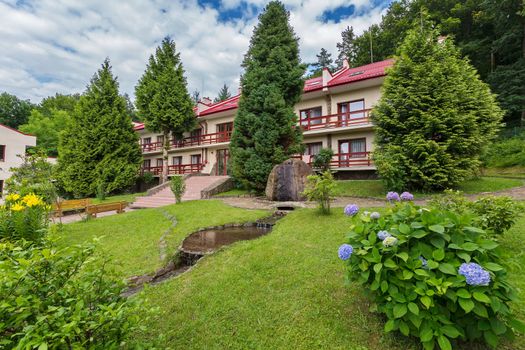 Decorative stone fountain, green shrubs with flowers and tall coniferous trees at the entrance of the hotel
