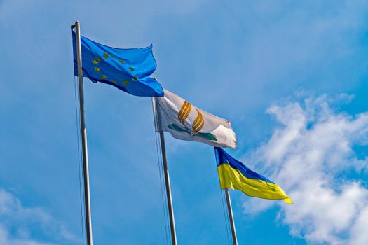 High flagpoles with flags of states developing under the gusts of wind against the background of a light blue sky with rare clouds.