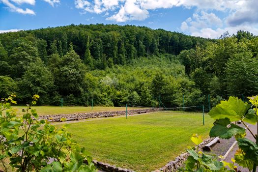 A volleyball court with green grass outdoors against the background of tall green trees. With benches for spare and a small wall around the site lined with stone.