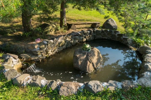memorable cheerful green toad on a rock in an artificial lake facing a stone under a tree