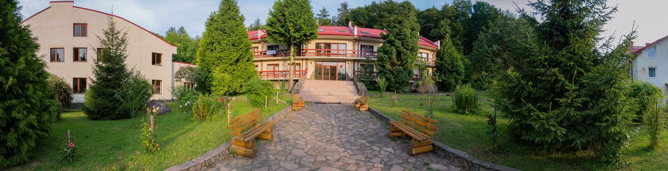 A chic panorama of the holiday complex located amidst green nature with a lawn and growing on it firs and a path laid out with a stone with standing benches leading to the entrance of the hotel.