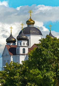 A majestic church with white walls and large domes with golden crosses glistening in the sun, standing among green trees against the sky covered with dense clouds.