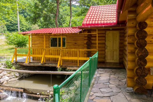 A small house built of wooden logs with a tiled roof with a stream flowing under it is located among the beautiful nature.