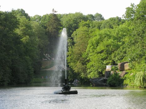 A high stream of a fountain beating in the middle of the water surface of the pond with tourists on the shore and an observation deck made on huge boulders.