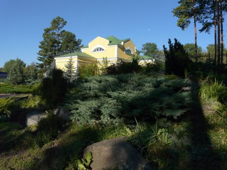 Large stones in a park lying among green lush bushes on the background of a house standing behind a fence of iron bars.