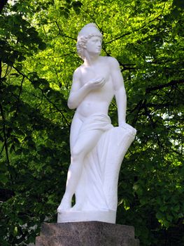 An elegant statue of a woman made of plaster. Standing on a pedestal among the green thickets of trees.