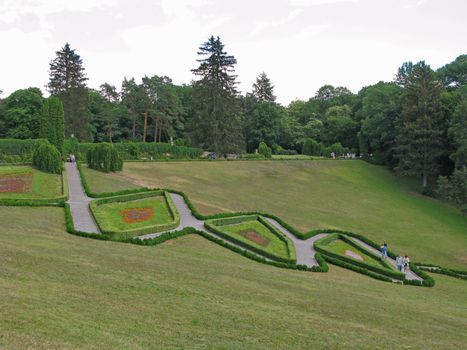 A beautiful landscape of a green lawn in a park on a slope with paths going between figured flower beds with people walking by them.