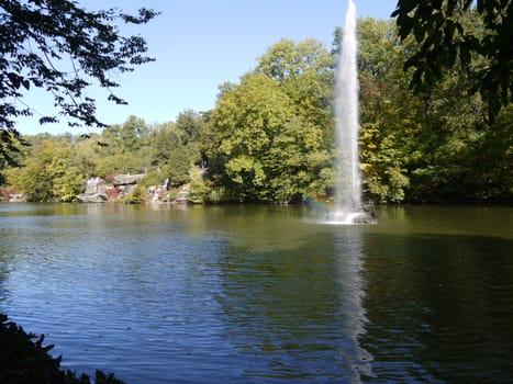 Tourists are resting on the shore of the reservoir with large stones lined on the shore among green trees and a fountain of water pouring in the middle.