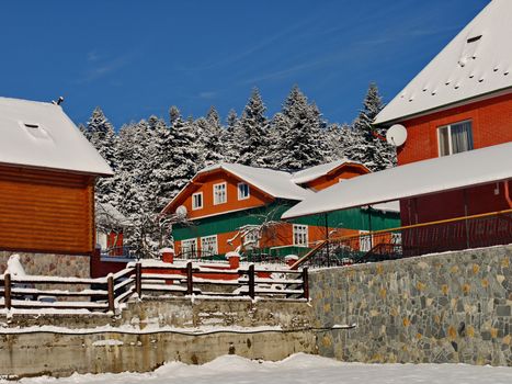 Several wooden houses covered with snow. In the background there is a pine forest